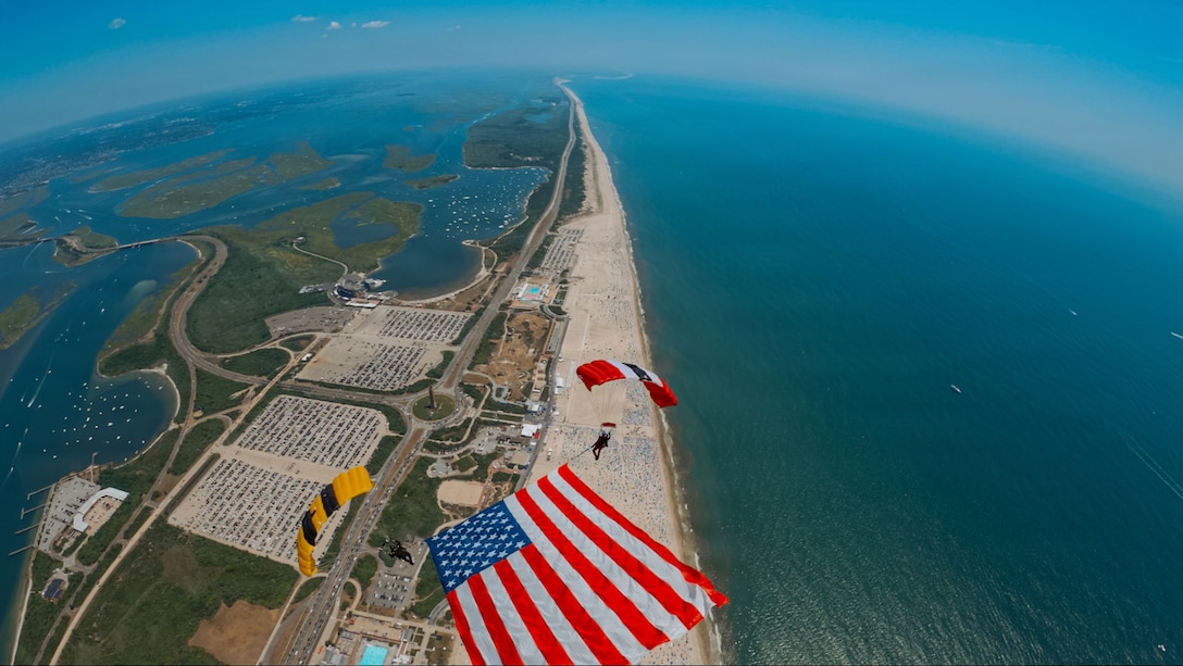 Service members display a large American flag while descending with parachutes over a strip of land surrounded by water during the day.