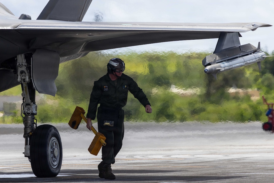 A Marine wearing a jumpsuit with a helmet and glasses carries blocks while walking under the wing of an aircraft during the day.
