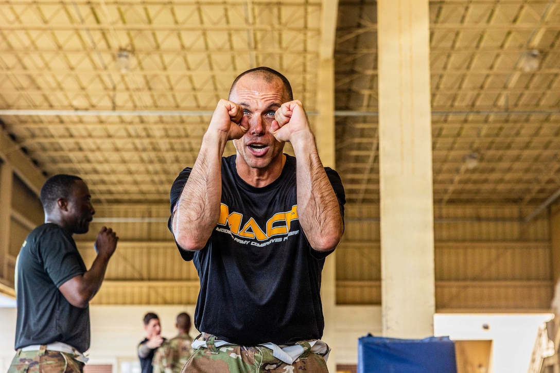 A soldier holds up two fists near their face while standing in a gymnasium with fellow soldiers training in the background.