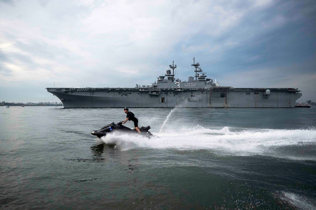 A Coast Guardsman makes a splash while riding a watercraft in front of a ship during the day.