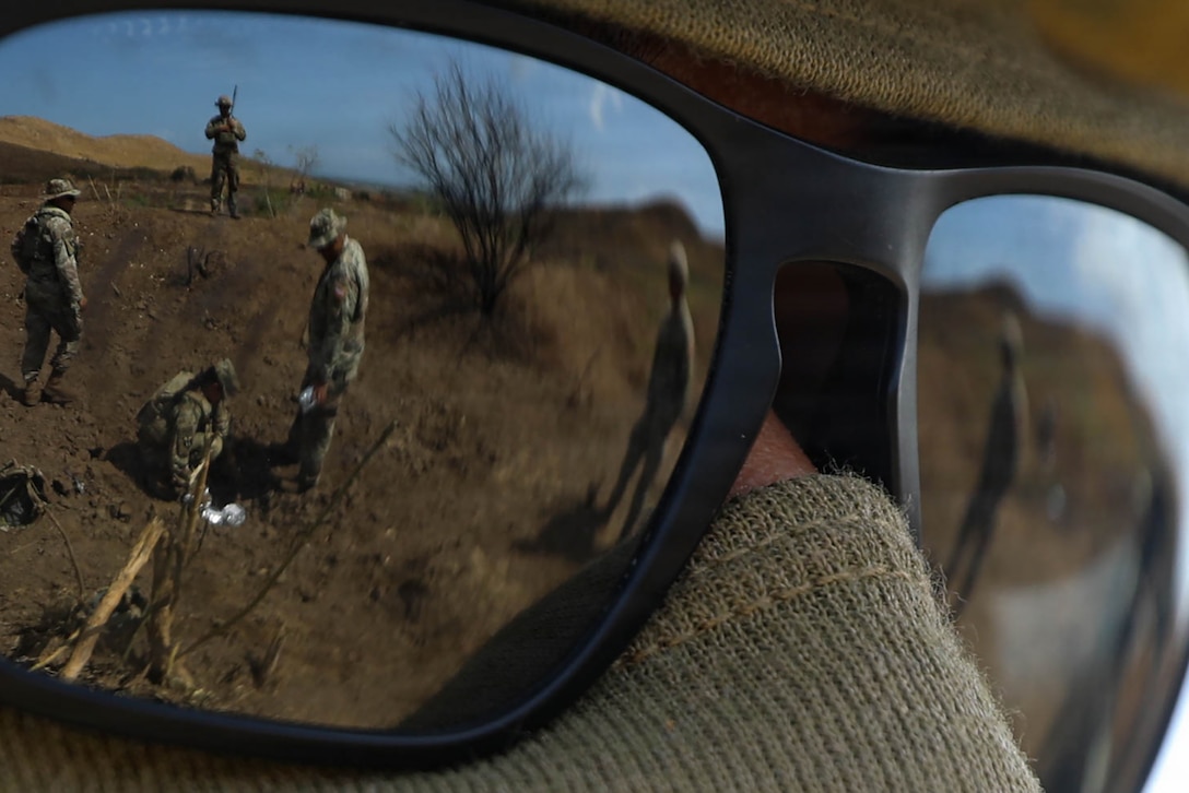 A soldier's view of other soldiers working on a dirt-covered hillside is reflected in sunglasses.