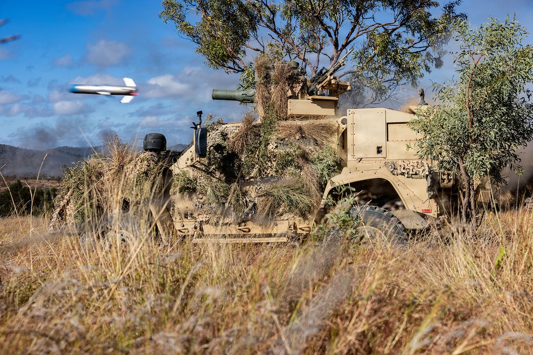 A missile is launched from a machine with leaves and twigs attached to it in a field. A tree and mountains are in the background.