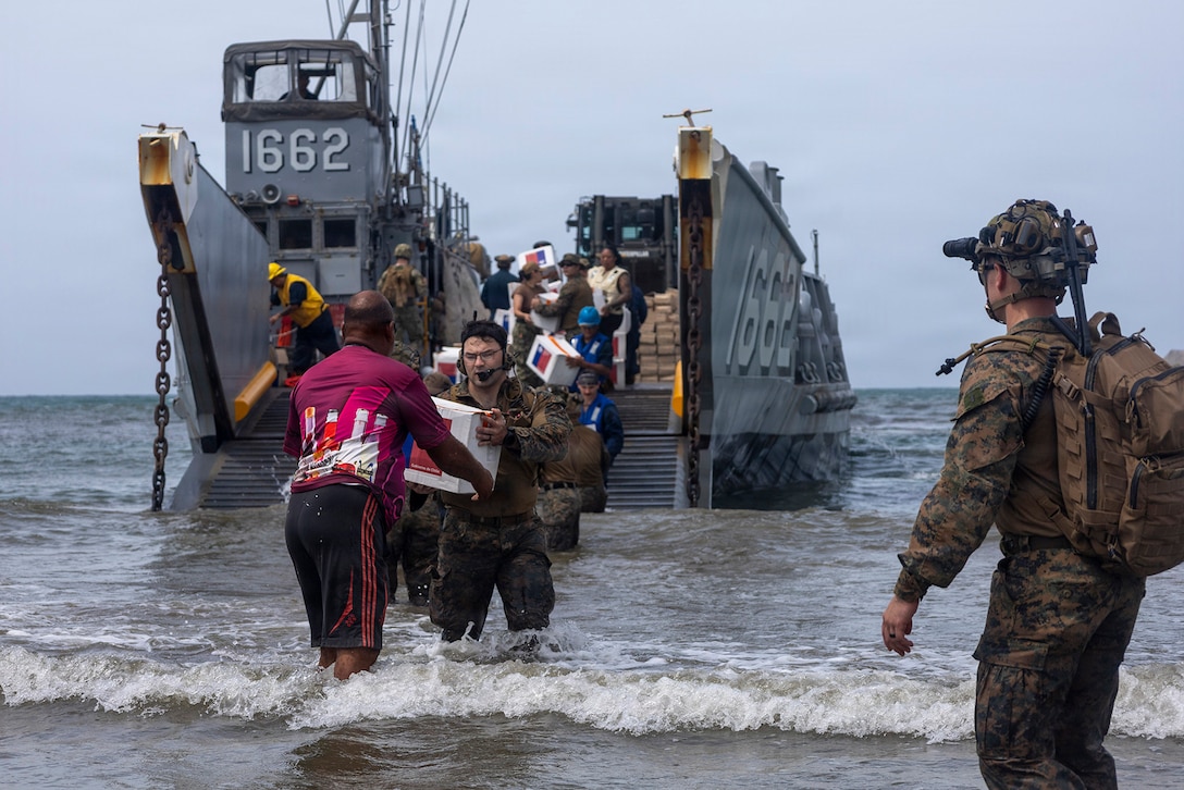 A U.S. service member hands a box to a civilian while standing on the shore as others behind them carry boxes off of a vessel. Another service member observes.