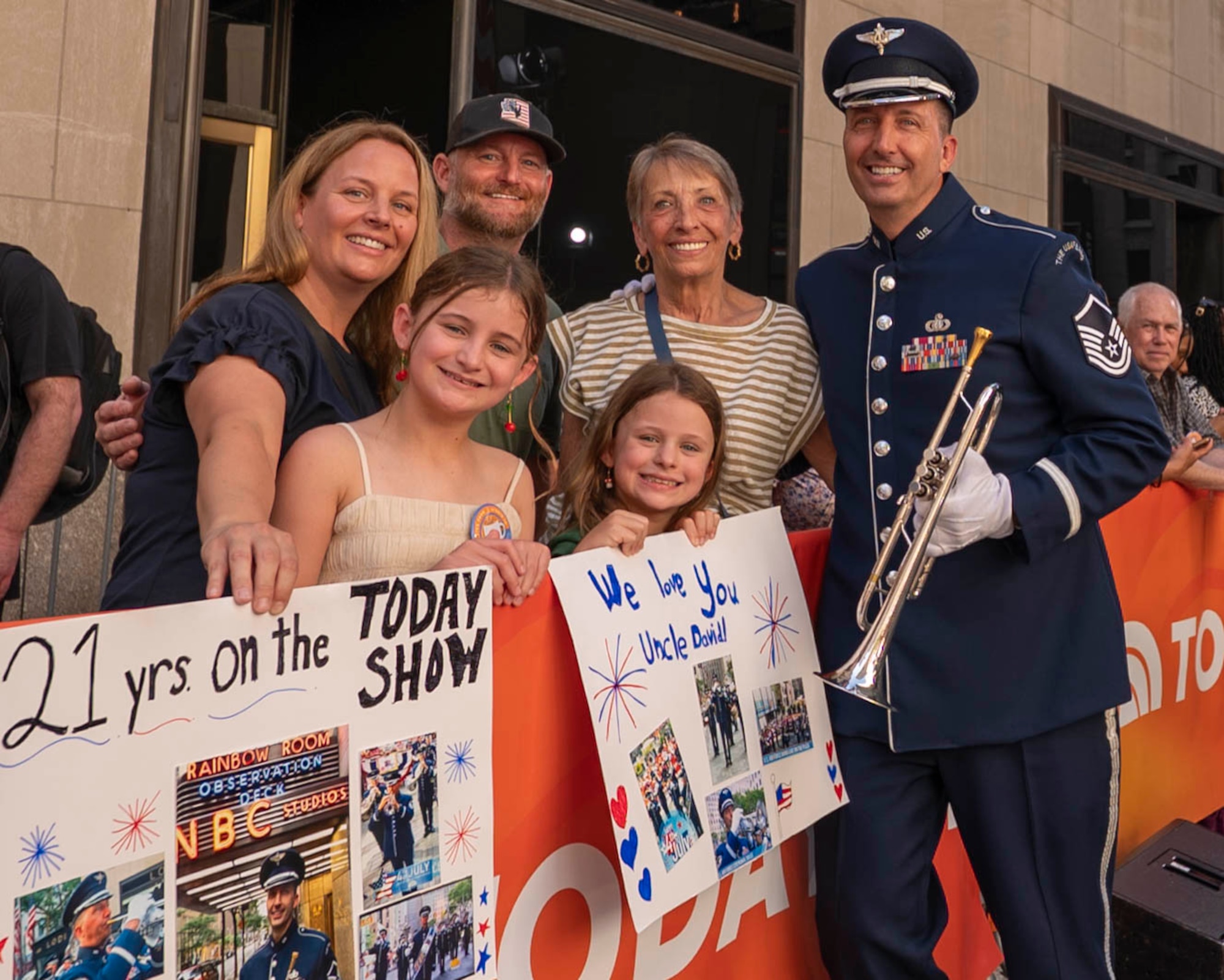 An Airman stands in ceremonial uniform holding a trumpet and smiliing next to relatives holding signs reading "21 years on the TODAY Show."