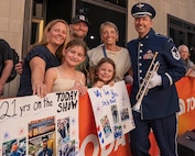 An Airman stands in ceremonial uniform holding a trumpet and smiliing next to relatives holding signs reading "21 years on the TODAY Show."