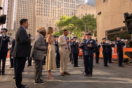 An Airman speaks on the set of the TODAY Show with the Air Force Band and city buildings behind her.