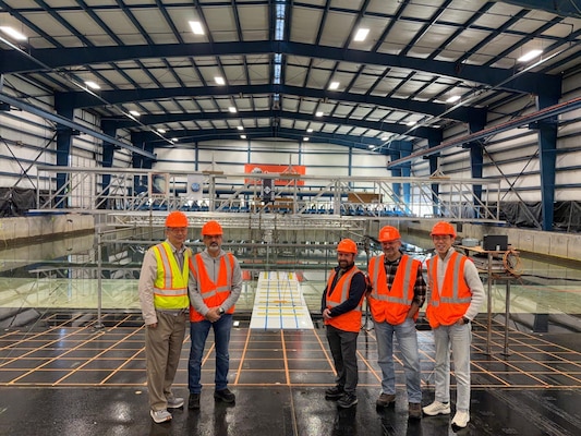 five people pose in warehouse-like structure with enormous wave pool behind them