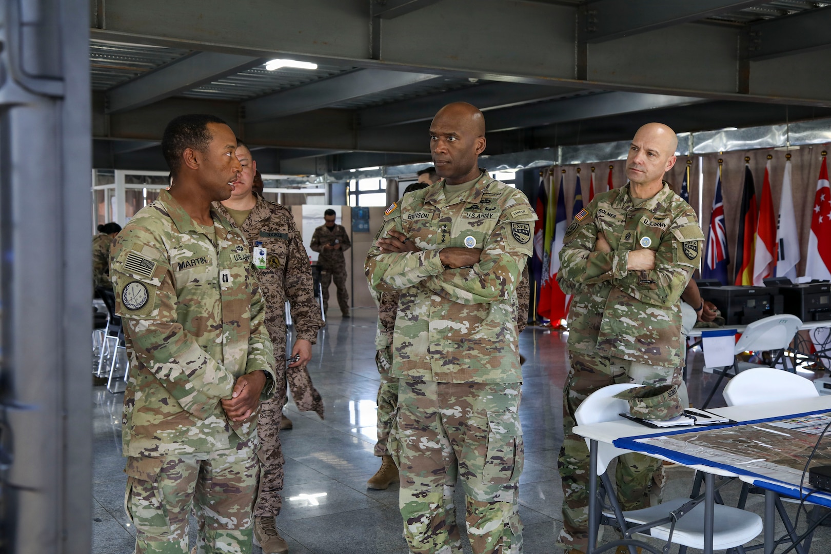 U.S. Army Capt. Makkonen Martin stands on the left in military uniform, gesturing as he briefs Gen. Xavier T. Brunson and Command Sgt. Maj. Robin M. Bolmer on operations outdoors at the Five Hills Training Area near Ulaanbaatar, Mongolia, during exercise Khaan Quest 2026.
