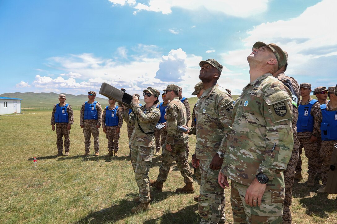 U.S. Army Staff Sgt. Alexis Caratti, a military police officer, stands in the center holding a drone buster device as she demonstrates counter-drone operations to Gen. Xavier T. Brunson and Command Sgt. Maj. Robin M. Bolmer outdoors at the Five Hills Training Facility near Ulaanbaatar, Mongolia, during exercise Khaan Quest 2026.