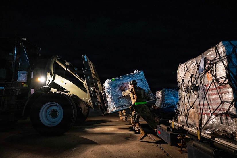 At night, airmen move a huge pallet of supplies onto a forklift from a larger pallet on the ground.