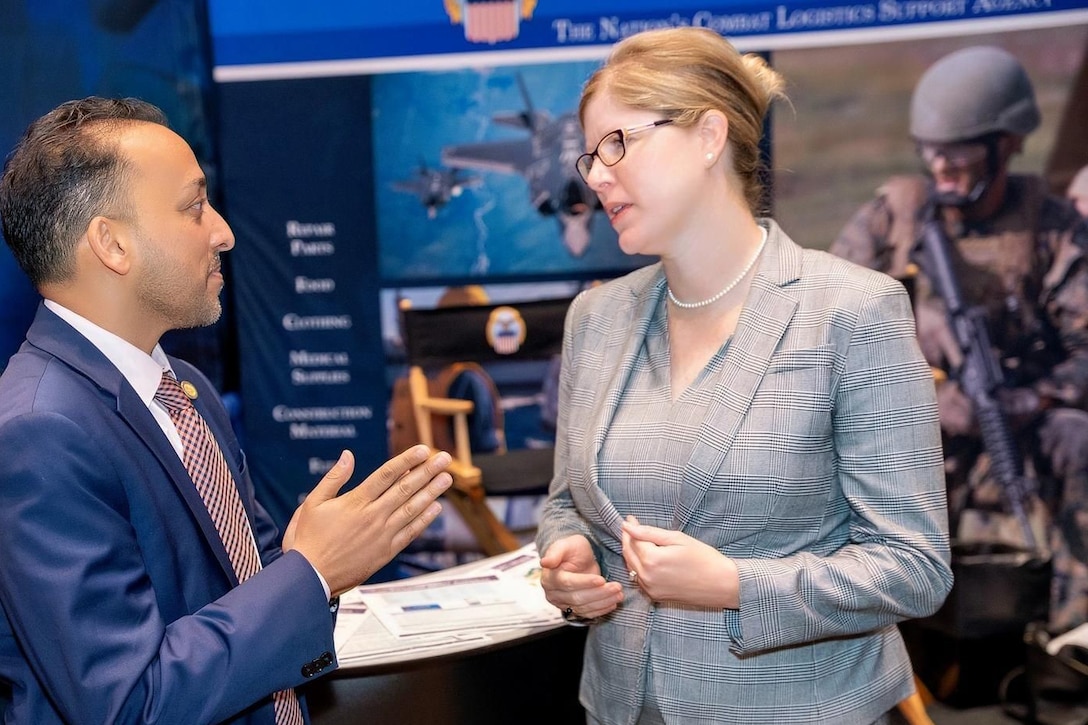 Two people in suits have a conversation in front of a convention floor booth.