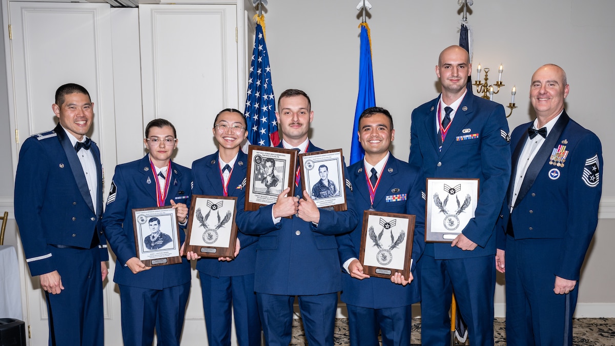 A group photo of men and women, wearing formal blue military uniforms. The five in the center hold award plaques