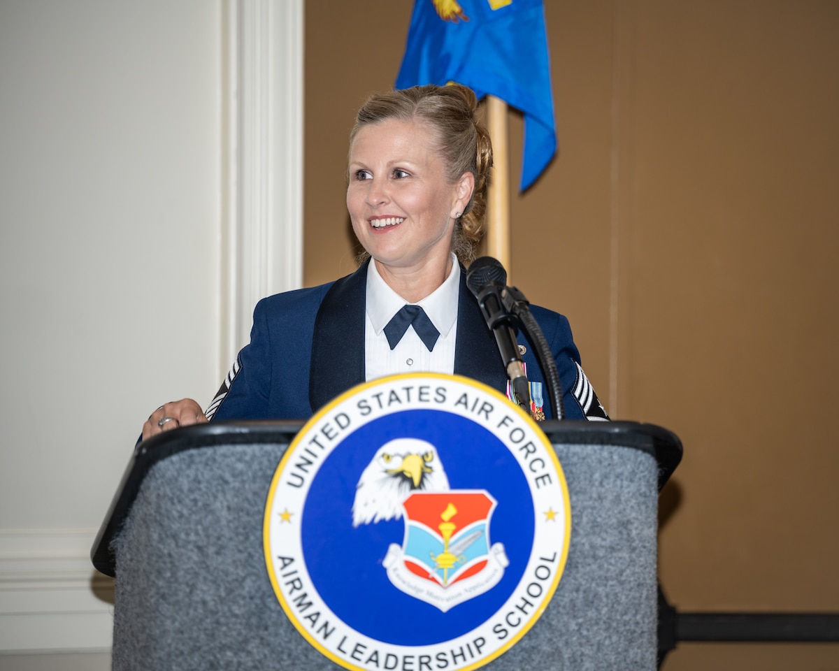 A blonde woman, wearing a blue formal military uniform, gives a speech behind a podium