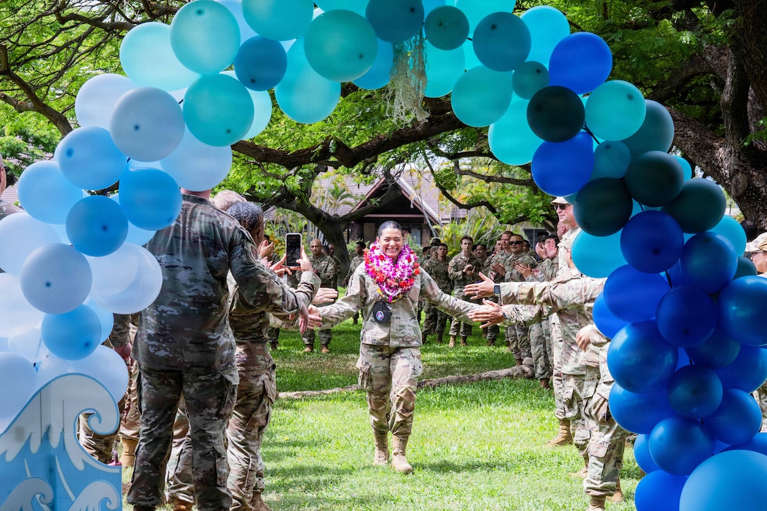 An airman wearing leis high-fives fellow service members standing in parallel lines in the grass while walking down the center toward a blue balloon arch.