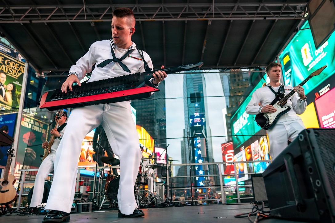 Sailors in white uniforms play instruments on a stage with tall city buildings in the background.