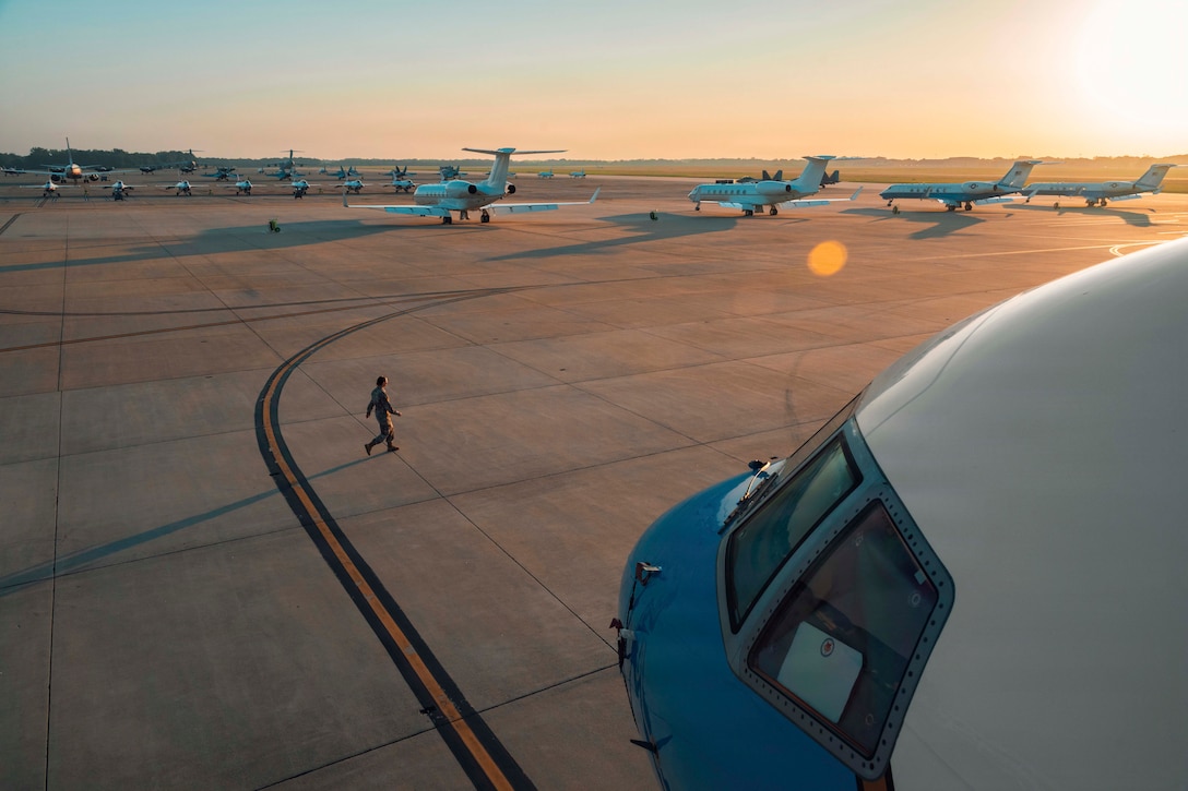 A service member walks near dozens of parked aircraft on a flight light as the sun shines in a pinkish sky to the right.