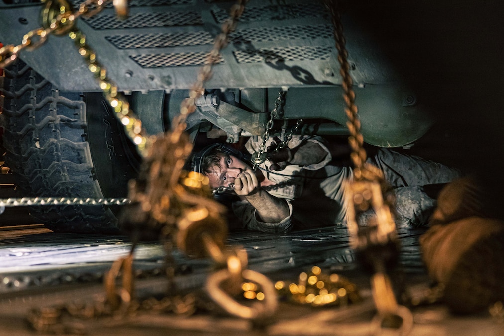 A Marine working under a military vehicle in the dark pulls a chain as seen through other chains in the foreground.