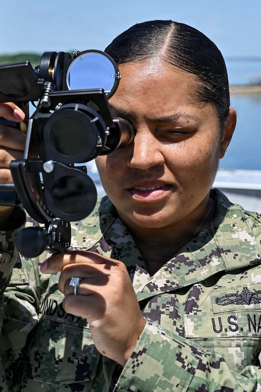 A woman wearing a camouflage military uniform peers into a military ocular device.
