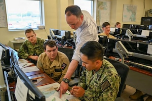 A man, wearing a white shirt, points to a map on a table as three other men in camouflage military uniforms look on.