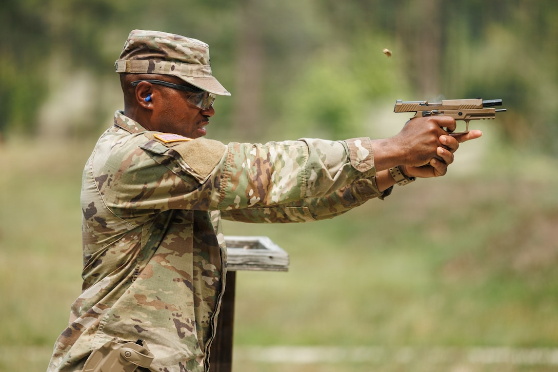 A soldier wearing a camouflage uniform fires a pistol on an outdoor range.