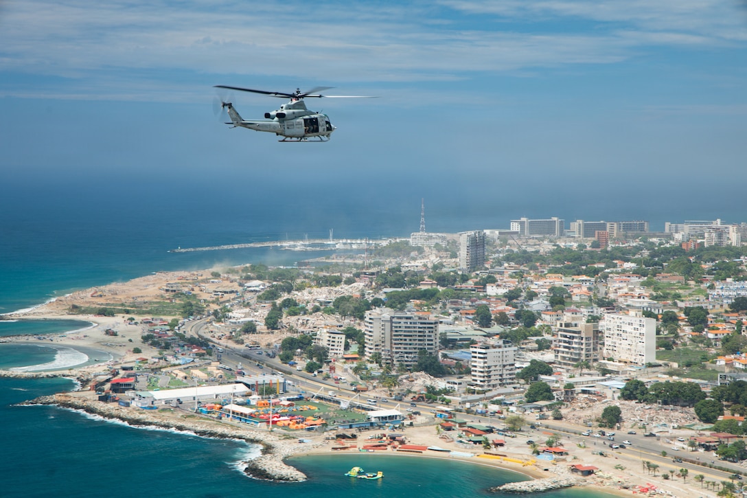 A helicopter flies above a body of water near a coastal city on a bright day.