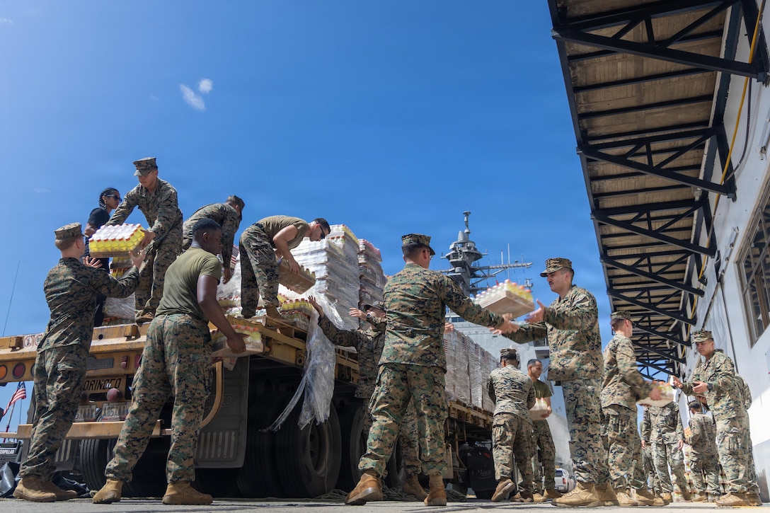 A group of about 15 Marines in camouflage uniforms unload cases of liquids from the back of a truck parked near a building in bright daylight.