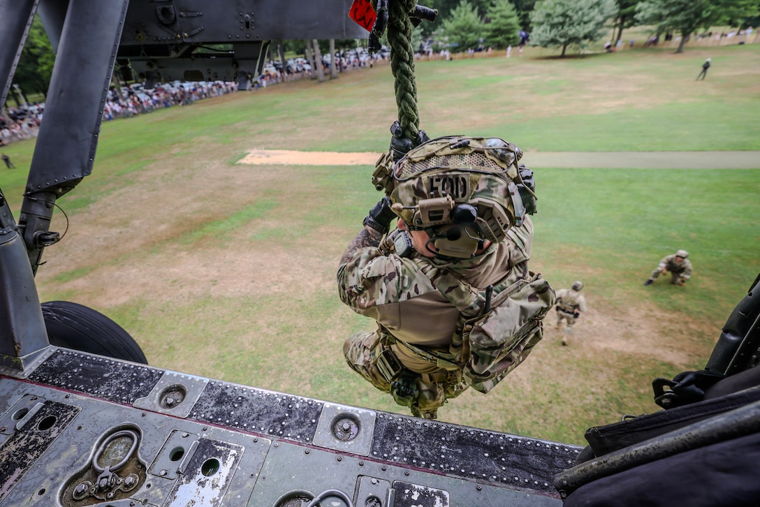 A sailor wearing a camouflage uniform and helmet slides down a rope from a helicopter hovering over a grassy field on a sunny day with a crowd of civilian spectators watching from a distance.