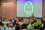 A uniformed man stands at the front of a room doing a brief