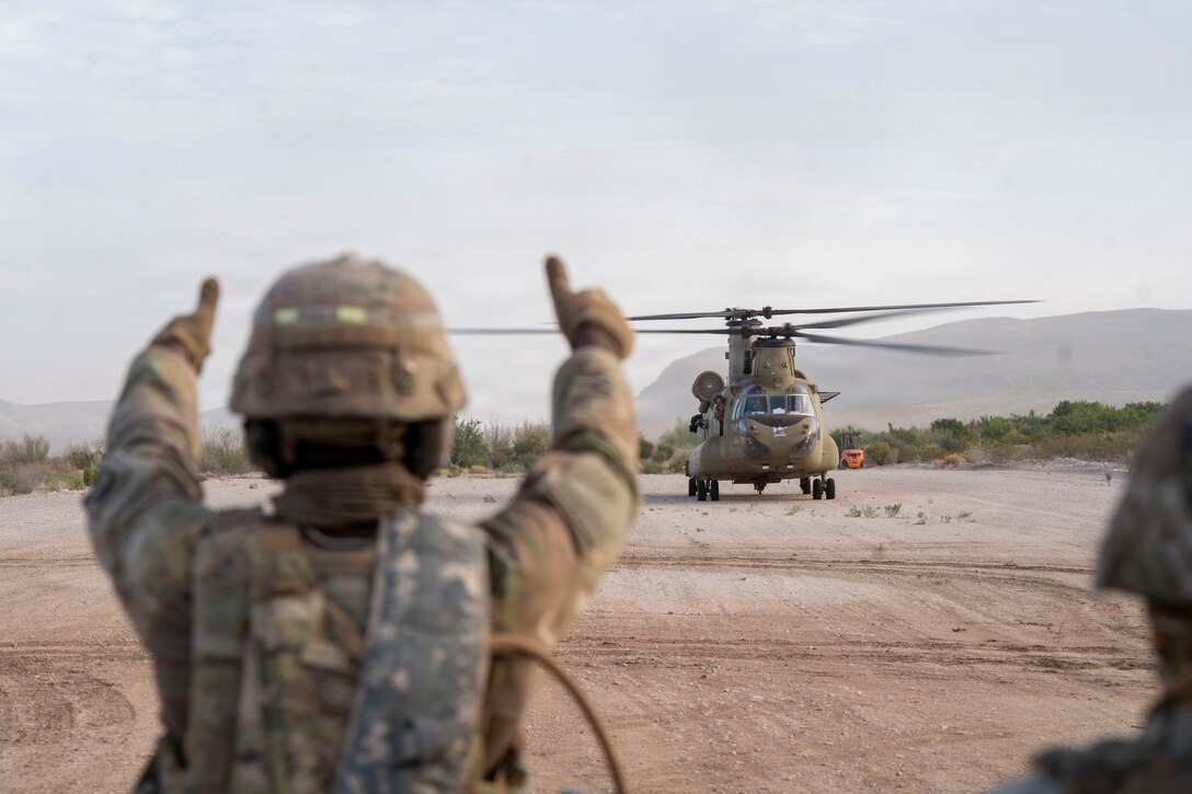 A soldier standing on a sandy lot gives two thumbs up to a helicopter about 100 yards away. Mountains are in the background.