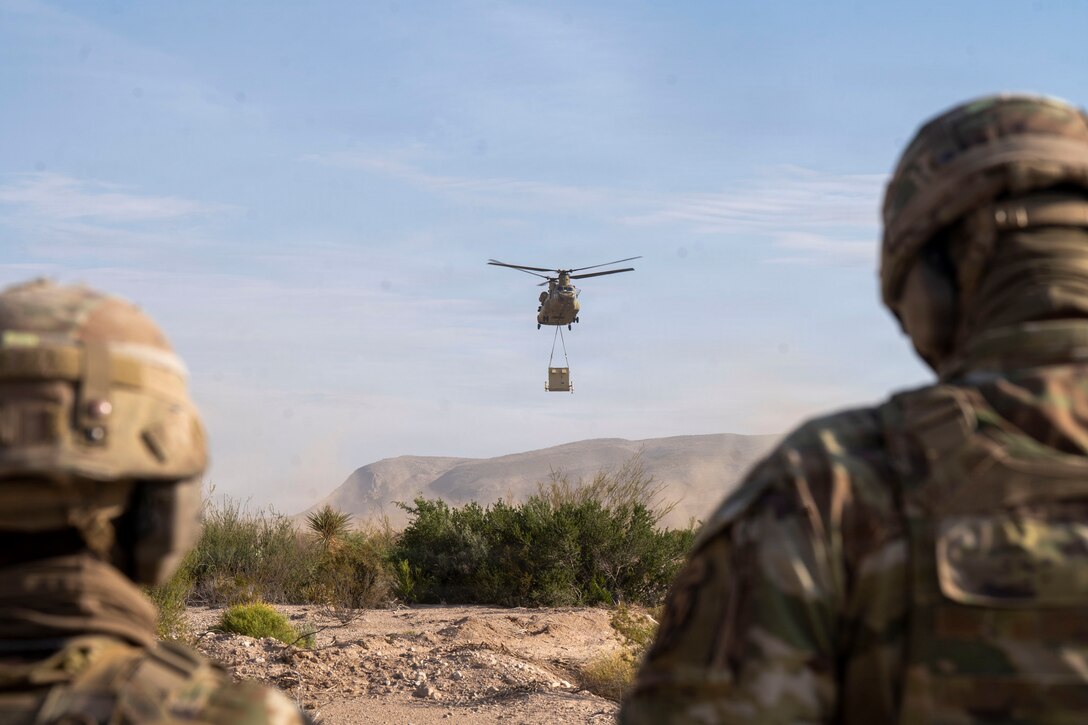 Two soldiers standing in the desert with their backs to the camera look up at a helicopter that has a large box hanging from it in a sling.
