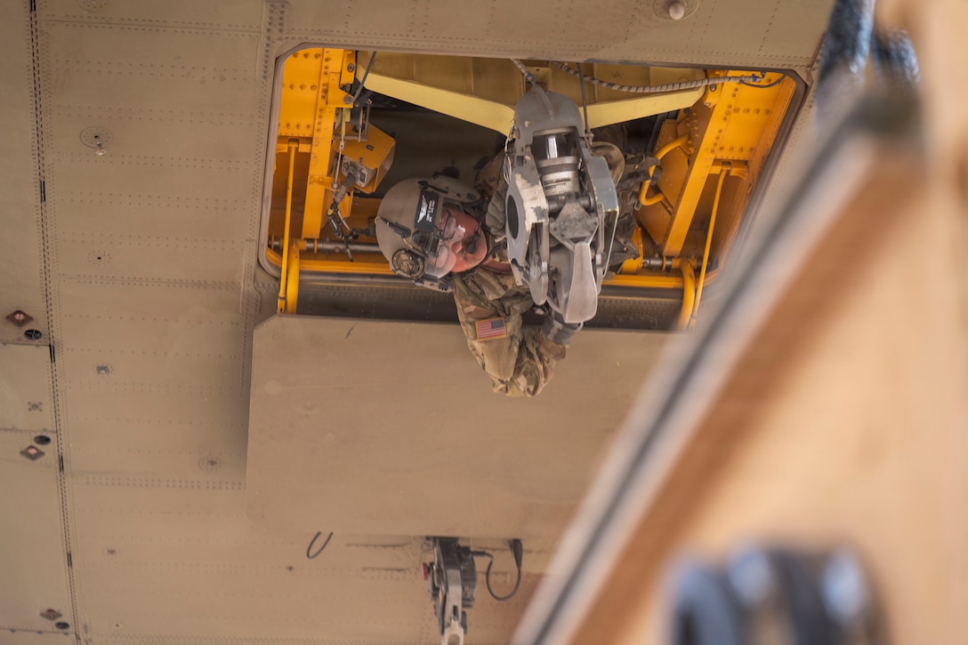 A soldier leans out of an opening in the bottom of a helicopter. One large hook attached to the helicopter is in front of him and another is behind him.