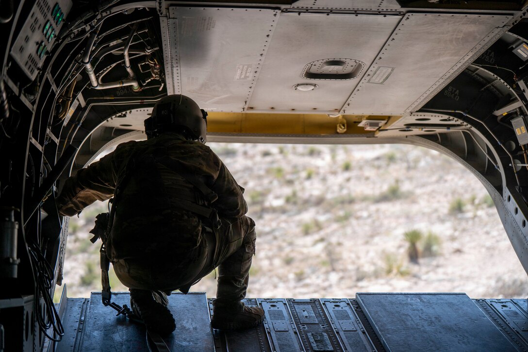 A soldier with their back toward the camera kneels on the cargo ramp of an airborne helicopter and looks at the desert terrain below.