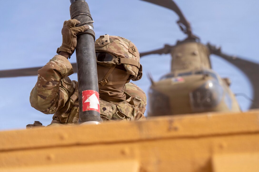 A soldier behind a yellow slab of metal holds onto a metal pipe that has a red sticker with a white arrow pointing up on it. A helicopter is in the background.