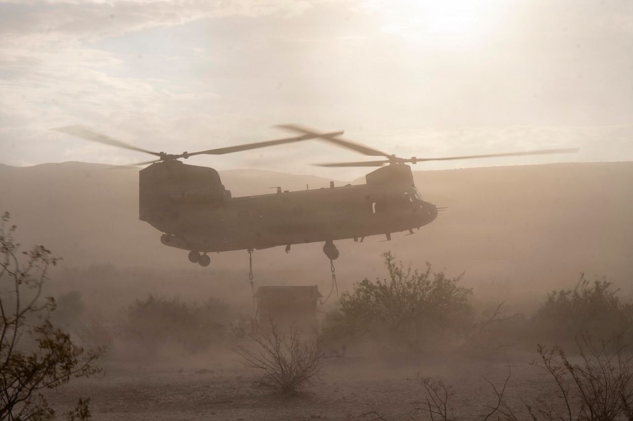 A large helicopter with a sling hanging from it hovers over a square piece of equipment in the dusty desert.