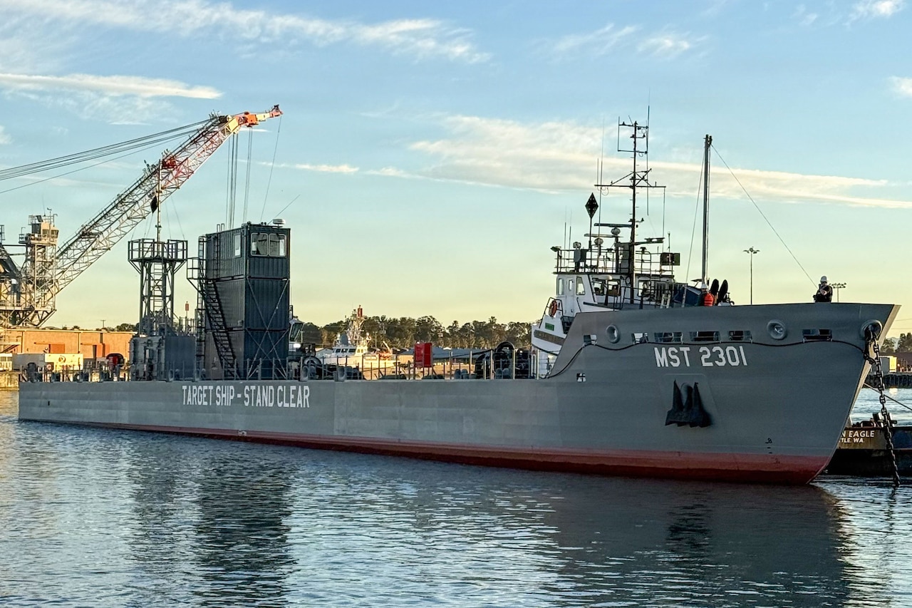 A large military ship is docked at a pier while a crane loads equipment onto the ship.