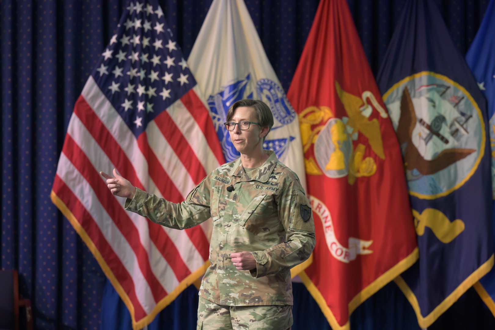 A female Army soldier stands on a stage with military flags in the background.