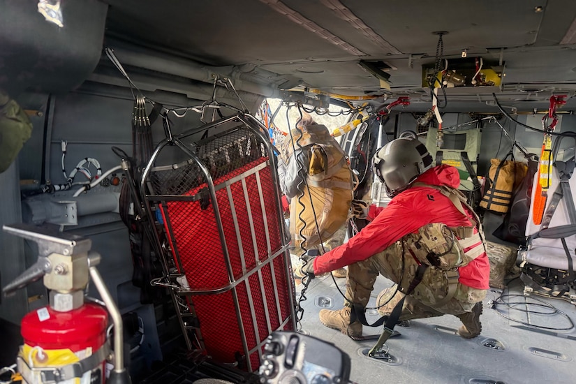 A person wearing a camouflage military uniform is strapped into a helicopter while pressing a controller to lift an attached harness as another person in similar attire guides a person inside the harness into the helicopter.