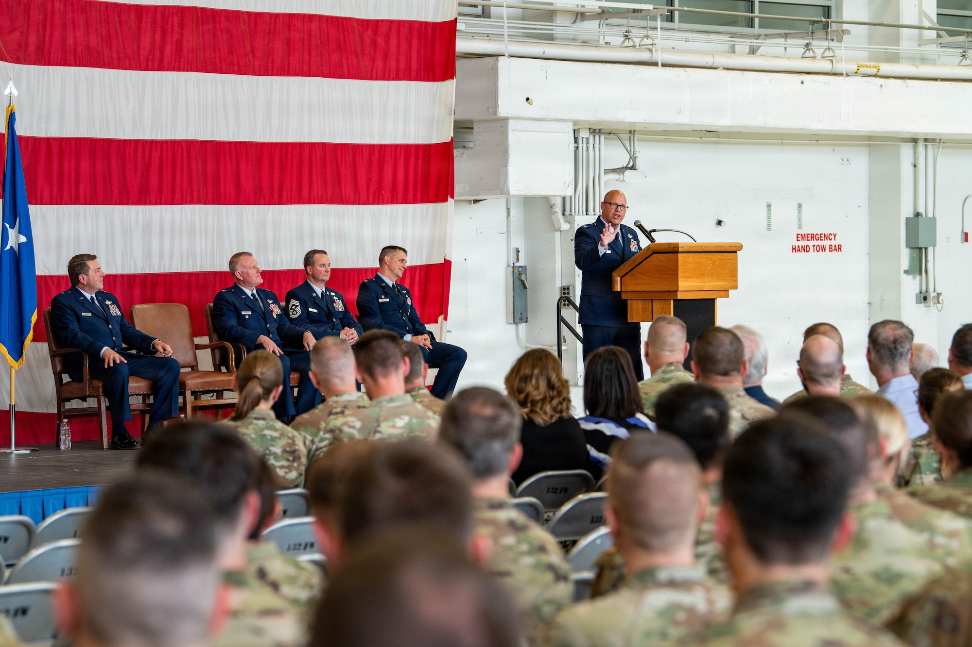 An Air Force officer in blue uniform stands on stage in front of large USA flag addressing seated military members.