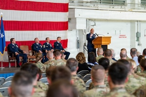 An Air Force officer in blue uniform stands on stage in front of large USA flag addressing seated military members.