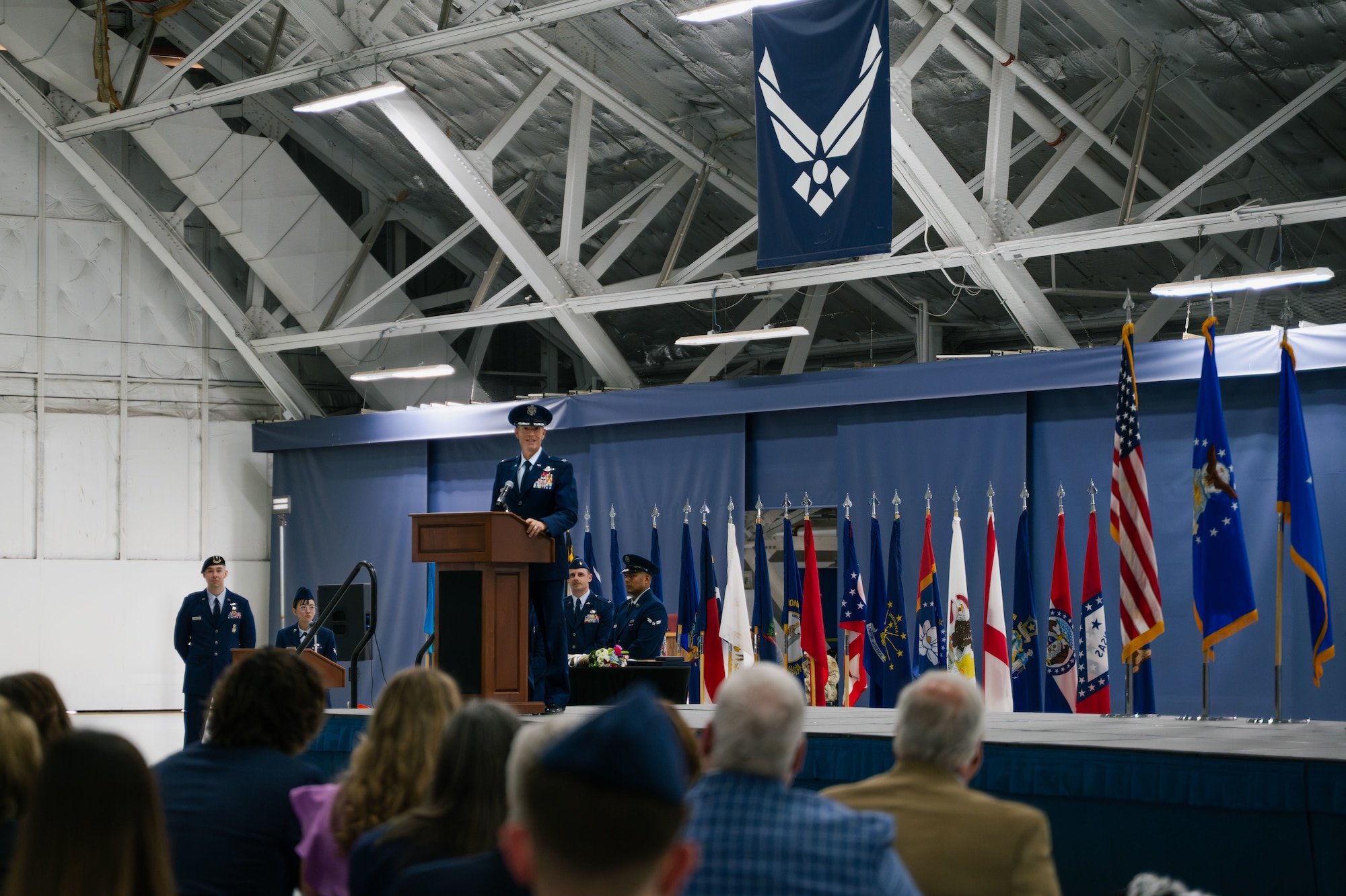 A man in U.S. Air Force service blues stands behind a podium and speaks to a large audience.