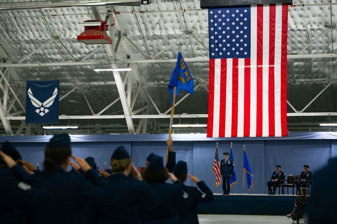 A man in U.S. Air Force service blues stands on a stage and receives a salute from U.S. Air Force service members in service blues.