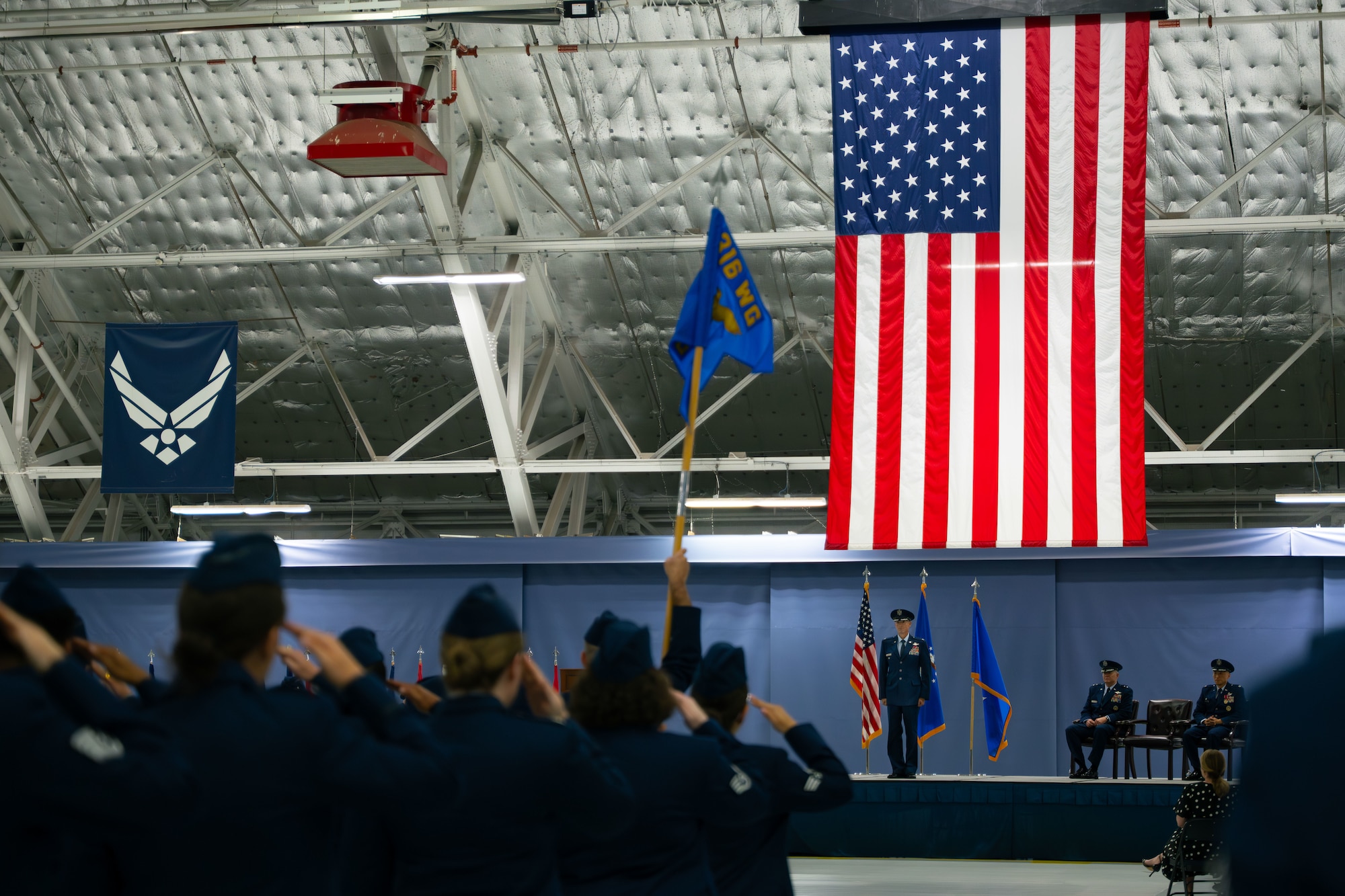 A man in U.S. Air Force service blues stands on a stage and receives a salute from U.S. Air Force service members in service blues.