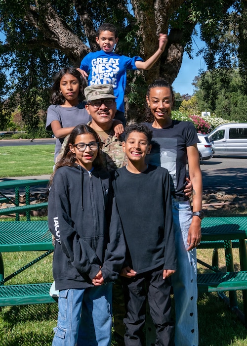 Family stands together in front of picnic table, youngest child jumps and smiles in background.