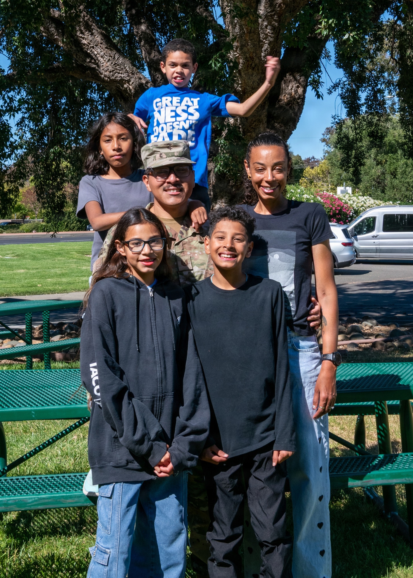 Family stands together in front of picnic table, youngest child jumps and smiles in background.