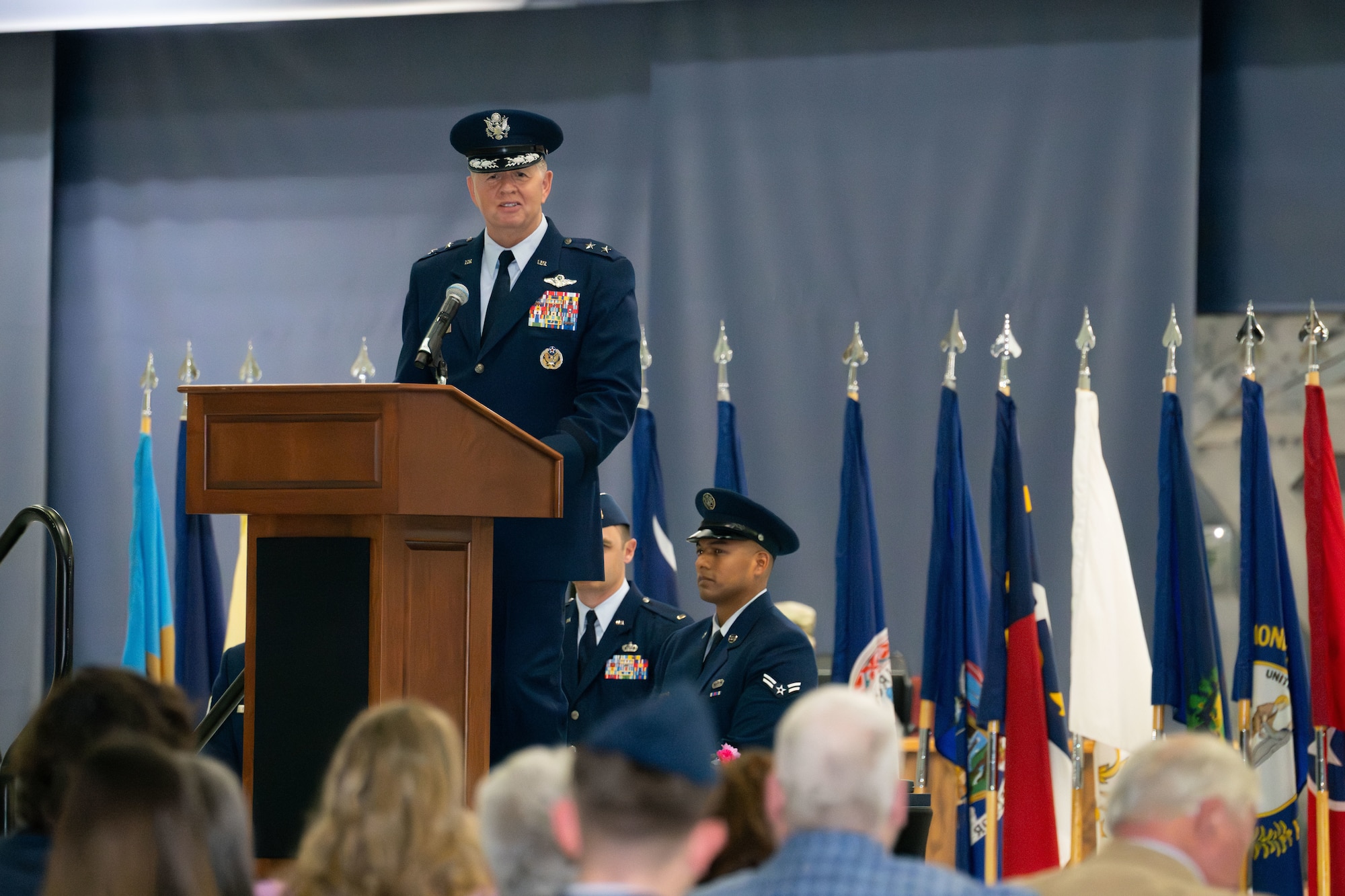 A man in U.S. Air Force service blues stands behind a podium and speaks to an audience.