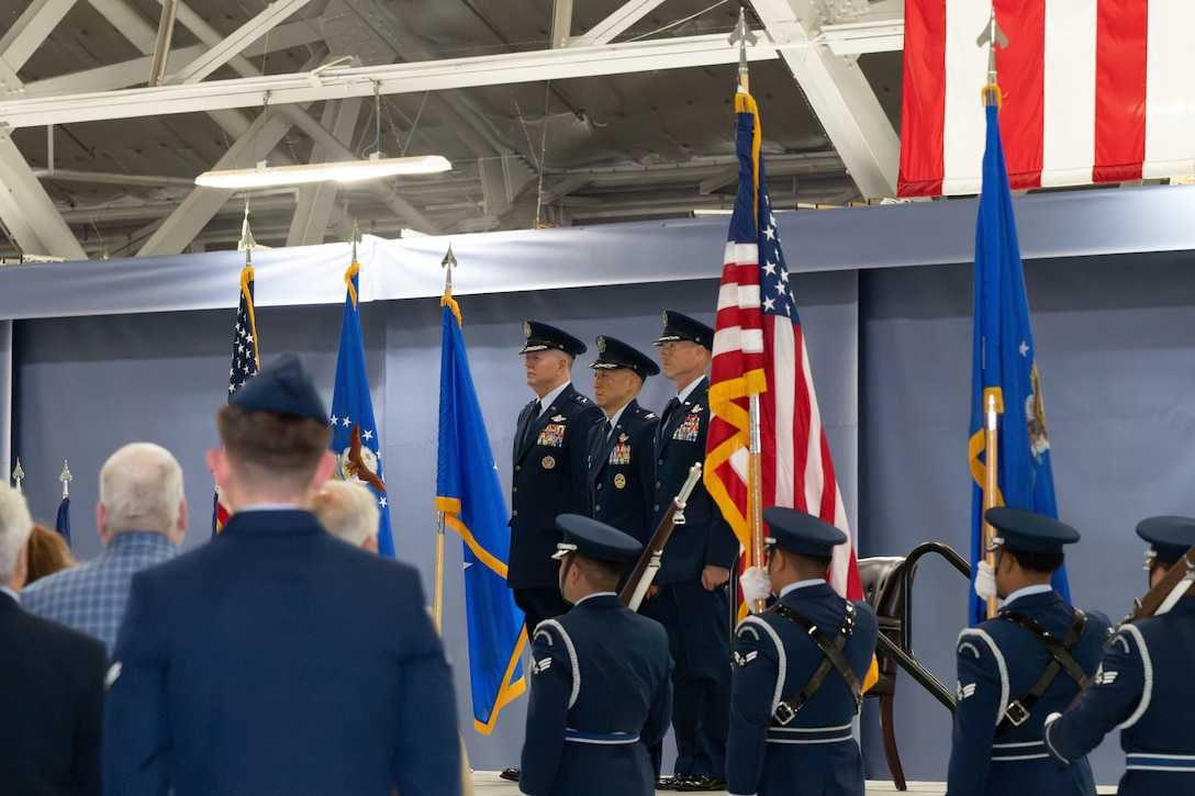 Three U.S. Air Force service members stand on a stage with their hands by their side as four U.S. service members holding flags pass them by.