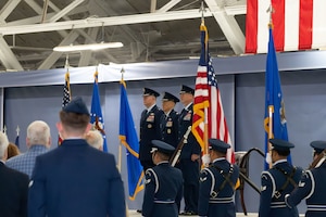Three U.S. Air Force service members stand on a stage with their hands by their side as four U.S. service members holding flags pass them by.