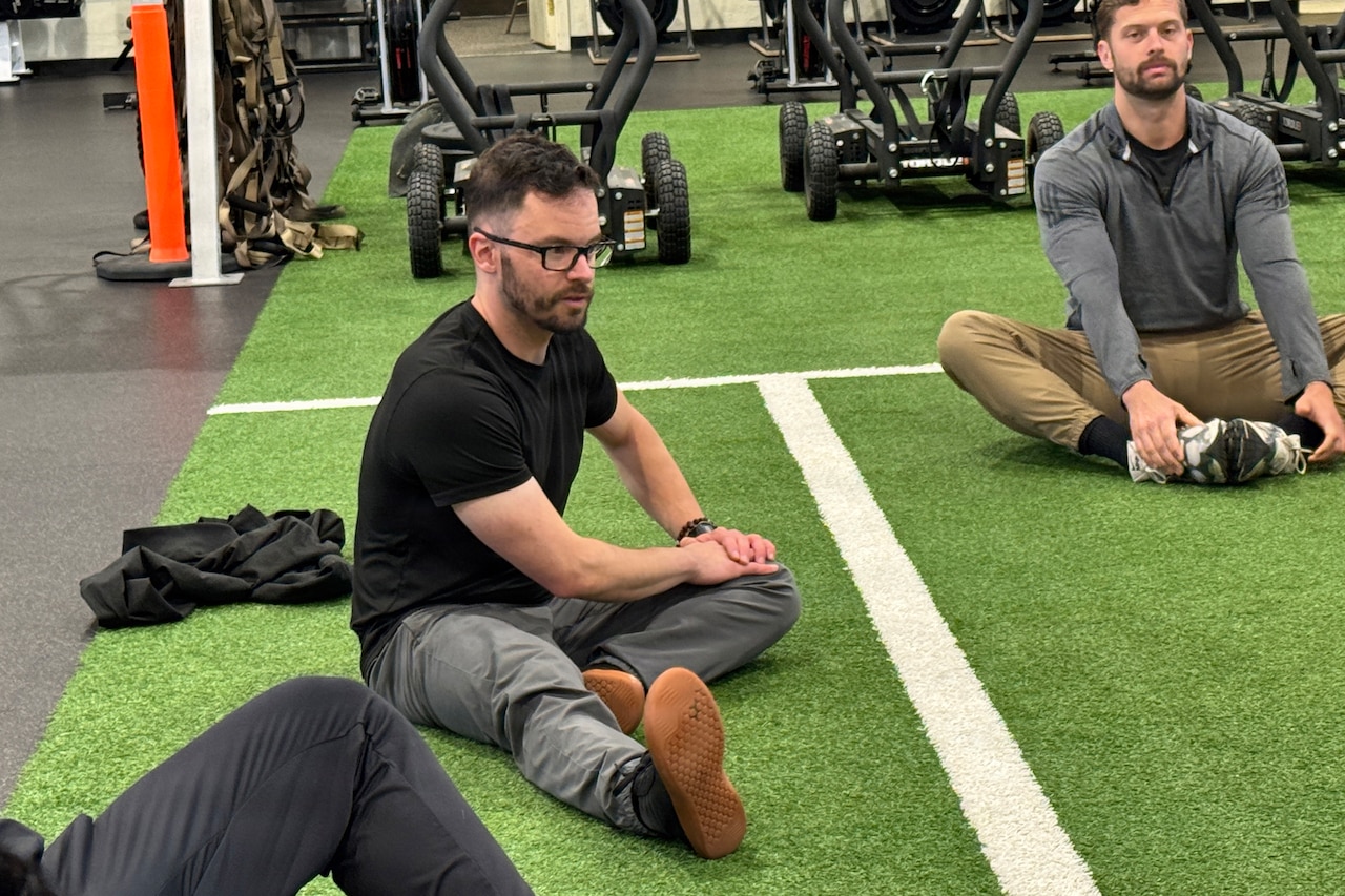 Two men sit on the floor of a gym and stretch their legs.