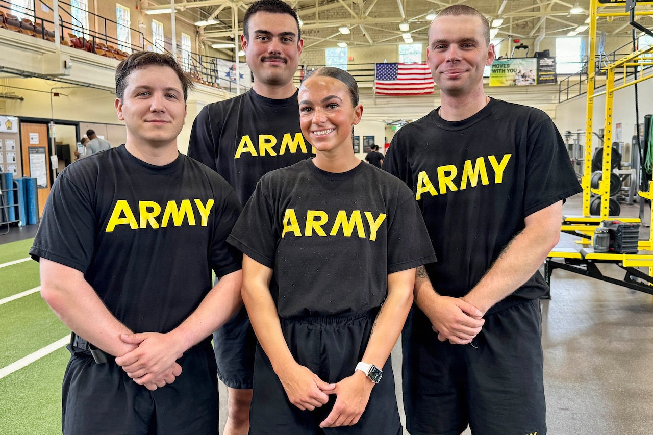 Three men and a woman dressed in black athletic gear stand together and smile. They are inside a gym. Behind them an American flag hangs from the ceiling.