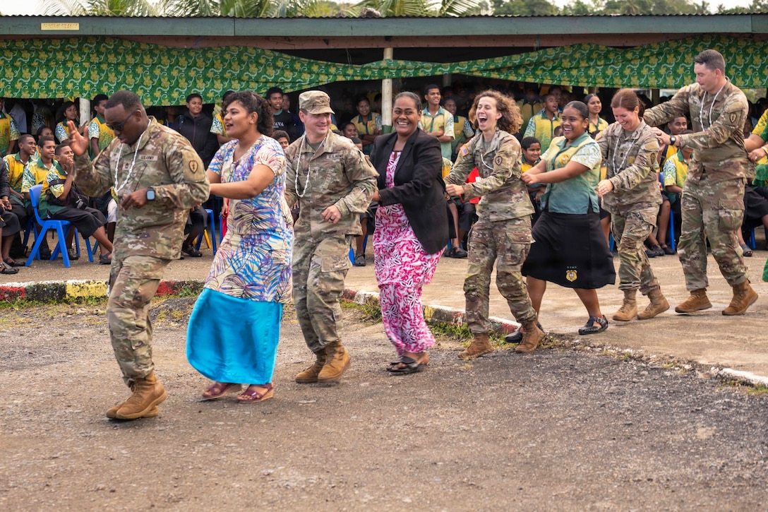 Soldiers and faculty members hold each other's waists while dancing in a line as spectators watch.